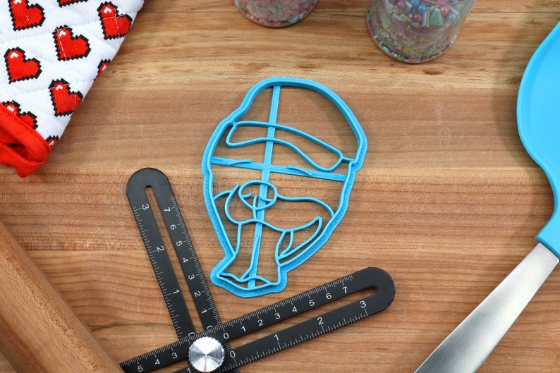 May include: Blue plastic cookie cutter shaped like a person wearing a hat. The cutter is on a wooden surface.