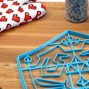 May include: A blue plastic cookie cutter shaped like a video game character's shield. The cookie cutter is on a wooden surface with a white oven mitt with red pixelated hearts in the background.
