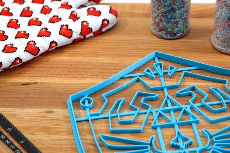May include: A blue plastic cookie cutter shaped like a video game character's shield. The cookie cutter is on a wooden surface with a white oven mitt with red pixelated hearts in the background.