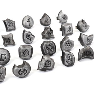 Puede incluir: Un conjunto de anillos plateados con varios s&iacute;mbolos grabados. Los anillos tienen un aspecto met&aacute;lico envejecido y est&aacute;n dispuestos sobre una superficie blanca. Los s&iacute;mbolos son intrincados y estilizados, sugiriendo un tema de fantas&iacute;a o juego.