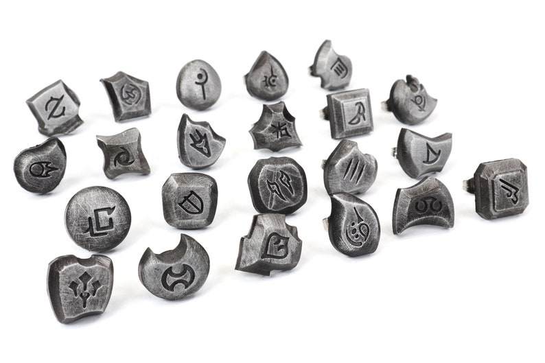 Puede incluir: Un conjunto de anillos plateados con varios s&iacute;mbolos grabados. Los anillos tienen un aspecto met&aacute;lico envejecido y est&aacute;n dispuestos sobre una superficie blanca. Los s&iacute;mbolos son intrincados y estilizados, sugiriendo un tema de fantas&iacute;a o juego.