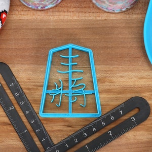 May include: A light blue plastic cookie cutter in the shape of a Japanese character, with a black ruler and a heart-patterned oven mitt on a wooden surface. The cookie cutter has the Japanese character "集" in the center.