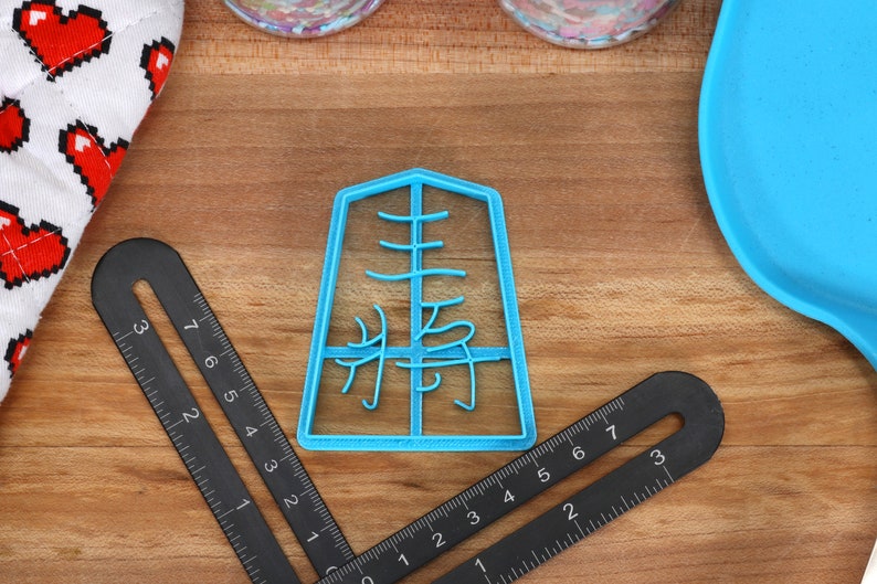 May include: A light blue plastic cookie cutter in the shape of a Japanese character, with a black ruler and a heart-patterned oven mitt on a wooden surface. The cookie cutter has the Japanese character "集" in the center.