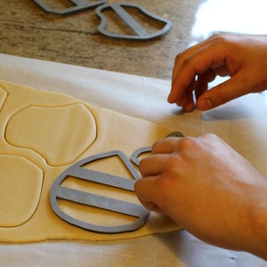 May include: A person using cookie cutters to cut out shapes from a sheet of cookie dough. The cutters are shaped like a bee and a honeycomb.