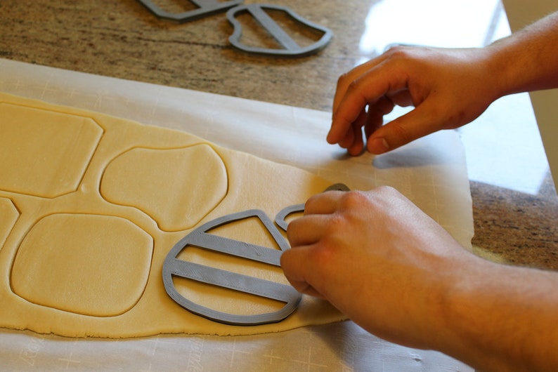 May include: A person using cookie cutters to cut out shapes from a sheet of cookie dough. The cutters are shaped like a bee and a honeycomb.