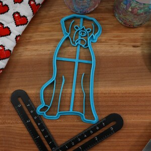 May include: Blue plastic cookie cutter shaped like a dog. The cutter is on a wooden surface with a ruler and a blue spatula.
