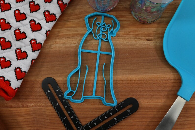 May include: Blue plastic cookie cutter shaped like a dog. The cutter is on a wooden surface with a ruler and a blue spatula.