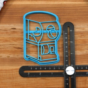 May include: Blue plastic cookie cutter shaped like a gumball machine with a clear window and a coin slot. The cutter is about 3 inches tall.