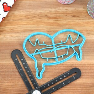 May include: A blue plastic cookie cutter shaped like a dripping heart. The cutter has a detailed design with multiple lines and curves.