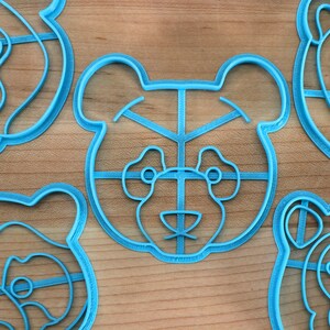 May include: Four blue plastic cookie cutters shaped like bear faces. Each cutter has a grid pattern on the face and a detailed outline of the bear's features.