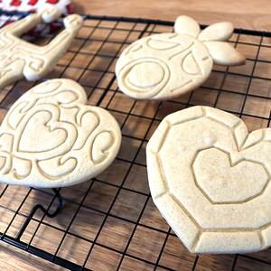 May include: Five cookies of different shapes and designs, including a heart, a pineapple, and a reindeer, are cooling on a black wire rack. The cookies are light brown and have intricate designs.