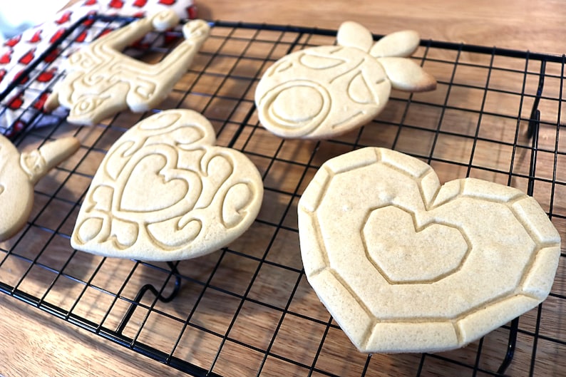 May include: Five cookies of different shapes and designs, including a heart, a pineapple, and a reindeer, are cooling on a black wire rack. The cookies are light brown and have intricate designs.