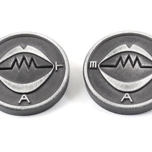 Może przedstawiać: Dwa srebrne metalowe żetony ze stylizowanym wzorem oka i literami "M", "A" i "F" wok&oacute;ł krawędzi.