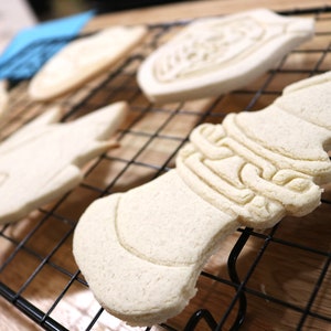 May include: A close-up of a wire rack with five unbaked sugar cookies. The cookies are in various shapes, including a fish, a bell, and a heart. The cookies are all white and have a smooth surface.