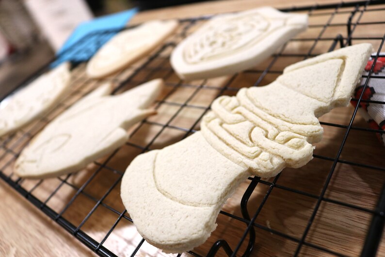 May include: A close-up of a wire rack with five unbaked sugar cookies. The cookies are in various shapes, including a fish, a bell, and a heart. The cookies are all white and have a smooth surface.