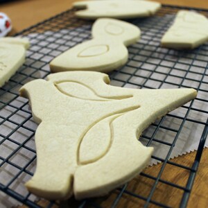 May include: Unbaked sugar cookies in various shapes, including a bottle, bird, and abstract designs, rest on a black wire cooling rack. The cookies are a light beige color, ready for baking and decorating.