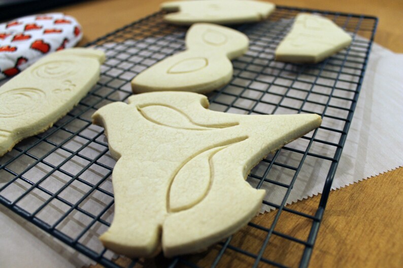 May include: Unbaked sugar cookies in various shapes, including a bottle, bird, and abstract designs, rest on a black wire cooling rack. The cookies are a light beige color, ready for baking and decorating.