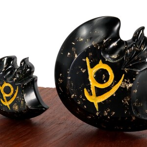May include: Two black obsidian crescent moon shaped sculptures with gold leaf accents and a stylized symbol in gold. The sculptures are on a wooden base.
