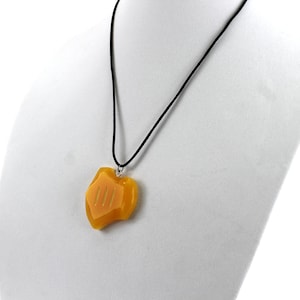May include: A yellow pendant necklace with a black cord. The pendant is shaped like a shield with three vertical lines on the front.