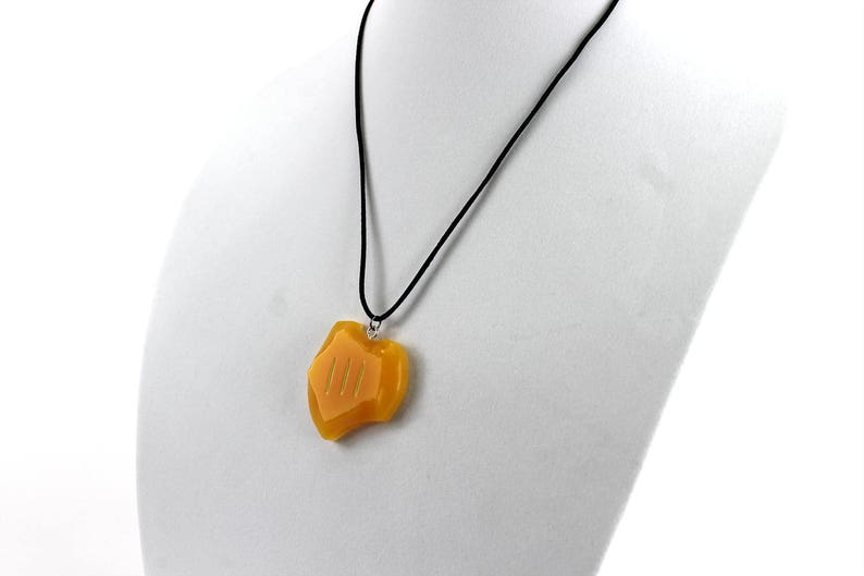 May include: A yellow pendant necklace with a black cord. The pendant is shaped like a shield with three vertical lines on the front.