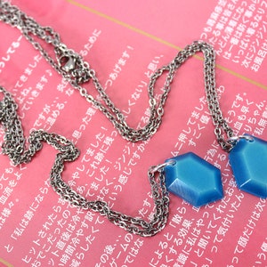 May include: Two silver chains with blue hexagonal pendants. The chains are layered and the pendants are hanging from the bottom of the chains.