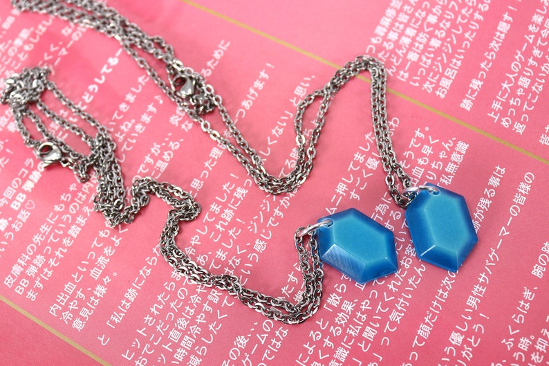 May include: Two silver chains with blue hexagonal pendants. The chains are layered and the pendants are hanging from the bottom of the chains.