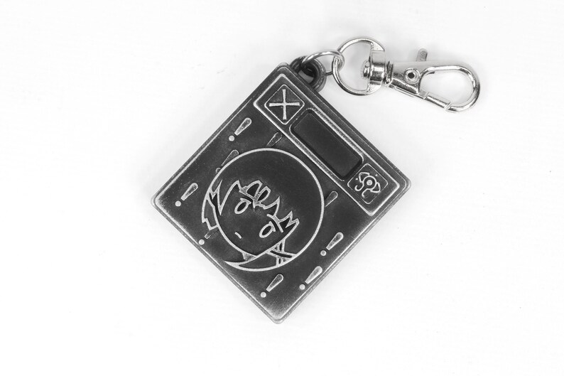 May include: A silver-colored keychain featuring a cartoon character with a surprised expression. The keychain has a small digital display and a button with a plus sign. The keychain is attached to a silver-colored metal ring.