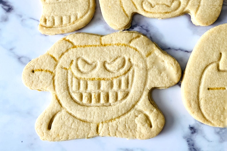 May include: A close-up of a sugar cookie decorated with a spooky face. The cookie is light brown and has a white icing outline. The face has two eyes, a wide smile, and sharp teeth.
