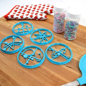 May include: A set of five blue cookie cutters with different geometric designs, a wooden rolling pin, a blue spatula, and two jars of colorful sprinkles on a wooden cutting board.