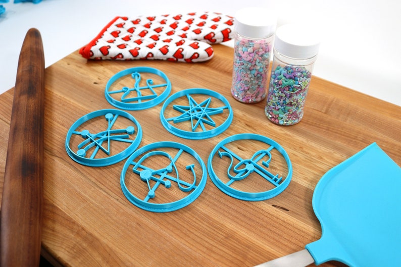 May include: A set of five blue cookie cutters with different geometric designs, a wooden rolling pin, a blue spatula, and two jars of colorful sprinkles on a wooden cutting board.