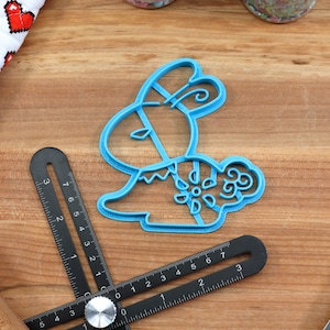 May include: A blue plastic cookie cutter shaped like a bunny rabbit with a floral design. The cutter is on a wooden surface.