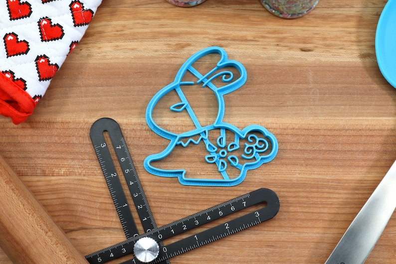 May include: A blue plastic cookie cutter shaped like a bunny rabbit with a floral design. The cutter is on a wooden surface.