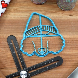 May include: Blue plastic cookie cutter shaped like a poodle wearing a hat. The cutter is on a wooden surface with a ruler and a rolling pin.