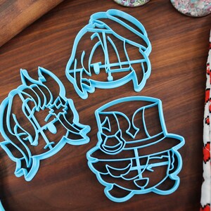 May include: Three blue plastic cookie cutters shaped like cartoon characters. The characters have large eyes, mouths, and hats.