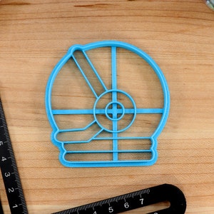 May include: A light blue plastic cookie cutter shaped like a snowman. The cutter has a round head with a circle for the face and two lines for the arms. The body is a rectangle with three lines for the buttons.