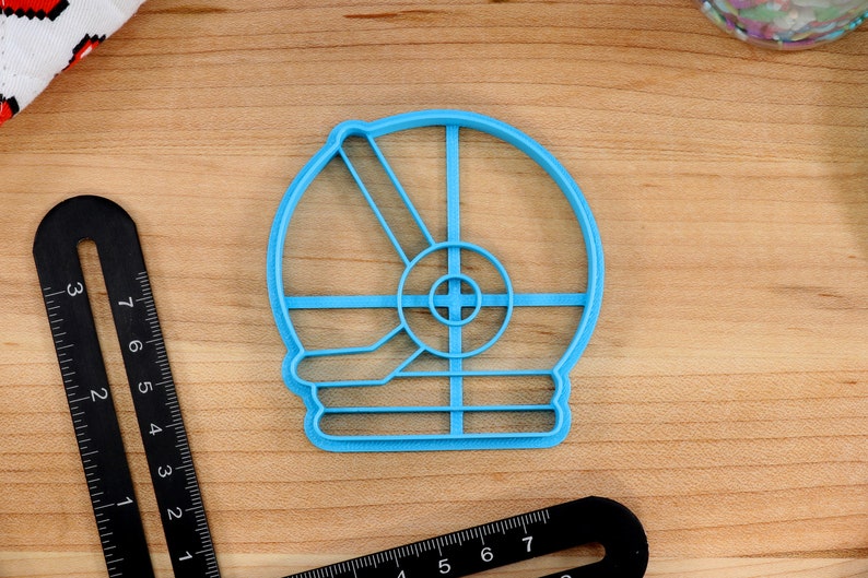 May include: A light blue plastic cookie cutter shaped like a snowman. The cutter has a round head with a circle for the face and two lines for the arms. The body is a rectangle with three lines for the buttons.