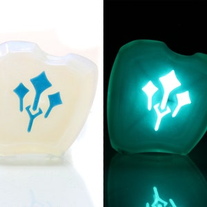 May include: A white, heart-shaped dice with a blue glow-in-the-dark symbol of three stylized leaves. The dice is shown in both normal and dark lighting.