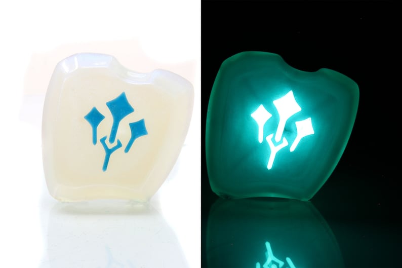 May include: A white, heart-shaped dice with a blue glow-in-the-dark symbol of three stylized leaves. The dice is shown in both normal and dark lighting.
