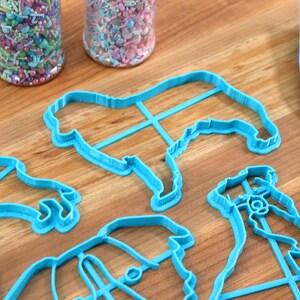 May include: Four blue plastic cookie cutters shaped like dogs, on a wooden surface with two jars of colorful sprinkles.