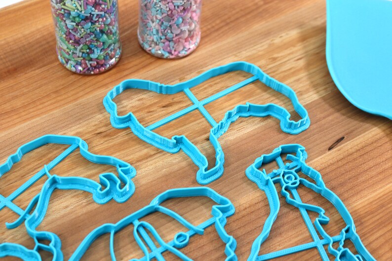 May include: Four blue plastic cookie cutters shaped like dogs, on a wooden surface with two jars of colorful sprinkles.