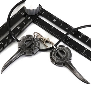 May include: Two silver-toned pendants with a circular design and a pointed tip hanging on a black cord. The pendants are on top of a black ruler with white markings.