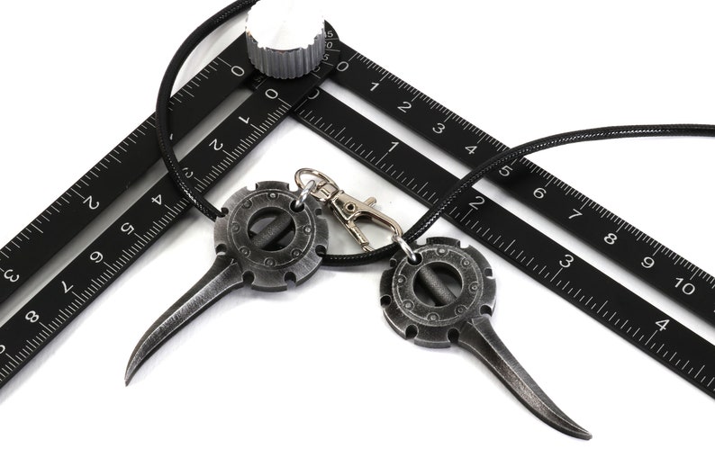May include: Two silver-toned pendants with a circular design and a pointed tip hanging on a black cord. The pendants are on top of a black ruler with white markings.
