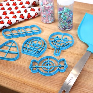 May include: A set of blue plastic Halloween cookie cutters in the shapes of a candy corn, a skull, a lollipop, a candy, and a row of five rectangles. The cutters are on a wooden surface with a rolling pin, a spatula, and two jars of sprinkles.