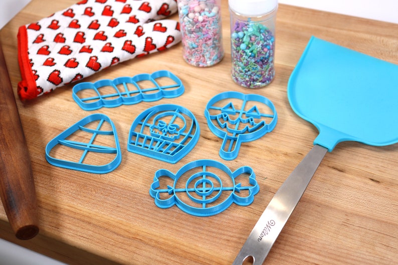 May include: A set of blue plastic Halloween cookie cutters in the shapes of a candy corn, a skull, a lollipop, a candy, and a row of five rectangles. The cutters are on a wooden surface with a rolling pin, a spatula, and two jars of sprinkles.