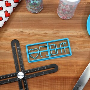 May include: Blue plastic cookie cutter with the words "I Fight" and a heart shape. The cutter is on a wooden surface with a ruler, rolling pin, and spatula.