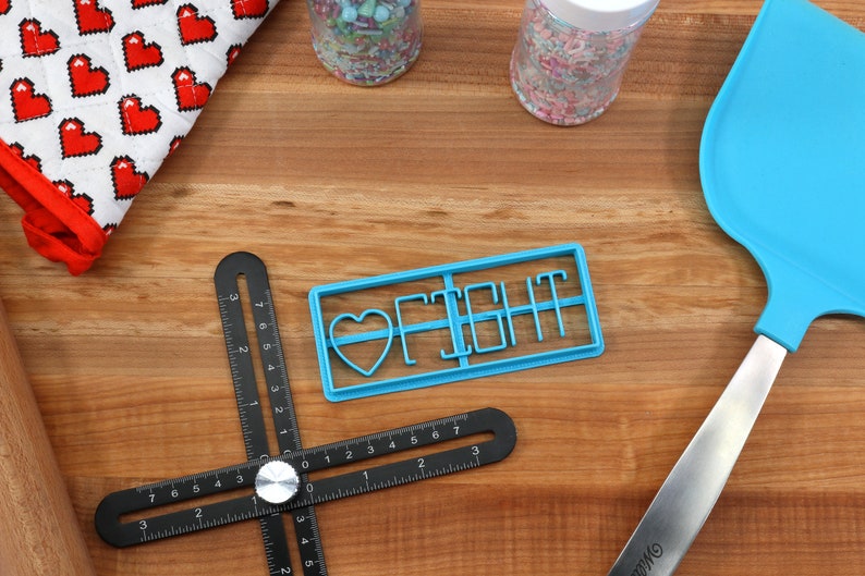 May include: Blue plastic cookie cutter with the words "I Fight" and a heart shape. The cutter is on a wooden surface with a ruler, rolling pin, and spatula.