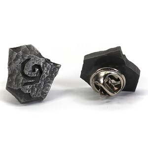 Puede incluir: Un pin de metal plateado con forma de roca irregular con un dise&ntilde;o de espiral estilizado. La parte posterior del pin tiene un cierre de pin est&aacute;ndar.