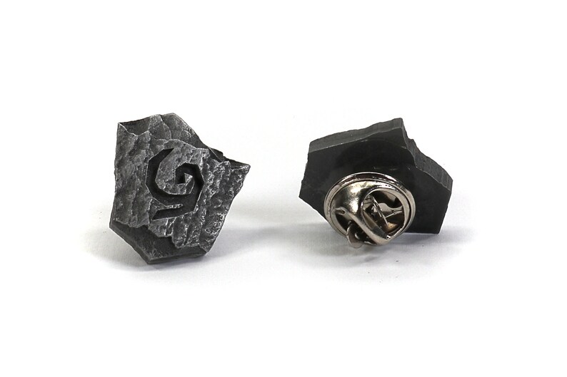 Puede incluir: Un pin de metal plateado con forma de roca irregular con un dise&ntilde;o de espiral estilizado. La parte posterior del pin tiene un cierre de pin est&aacute;ndar.