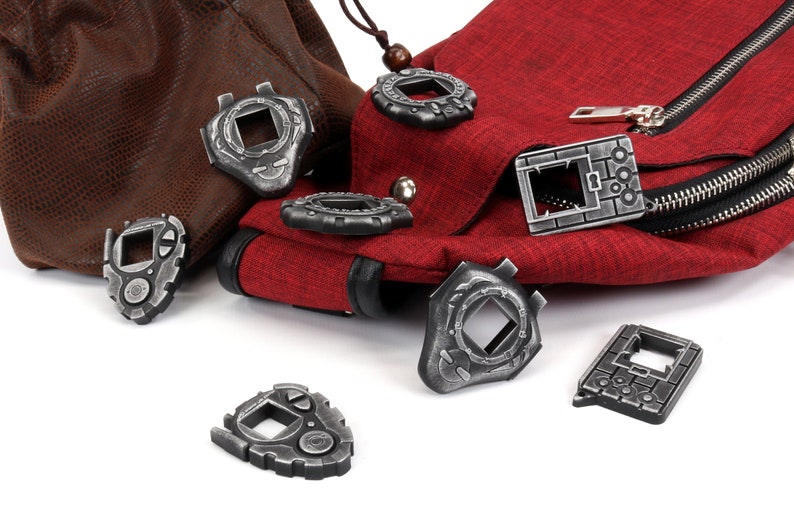 May include: A collection of eight silver-colored Digimon-style digital pet keychains. The keychains are scattered on a white surface, with two of them resting on a brown leather bag and a red fabric bag.