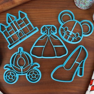 May include: A set of six blue plastic cookie cutters shaped like a castle, a dress, a shoe, a carriage, and two mice.